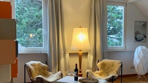A cozy room with two leather chairs, a white table, and a lamp. There are two windows with curtains, and a painting on the wall.