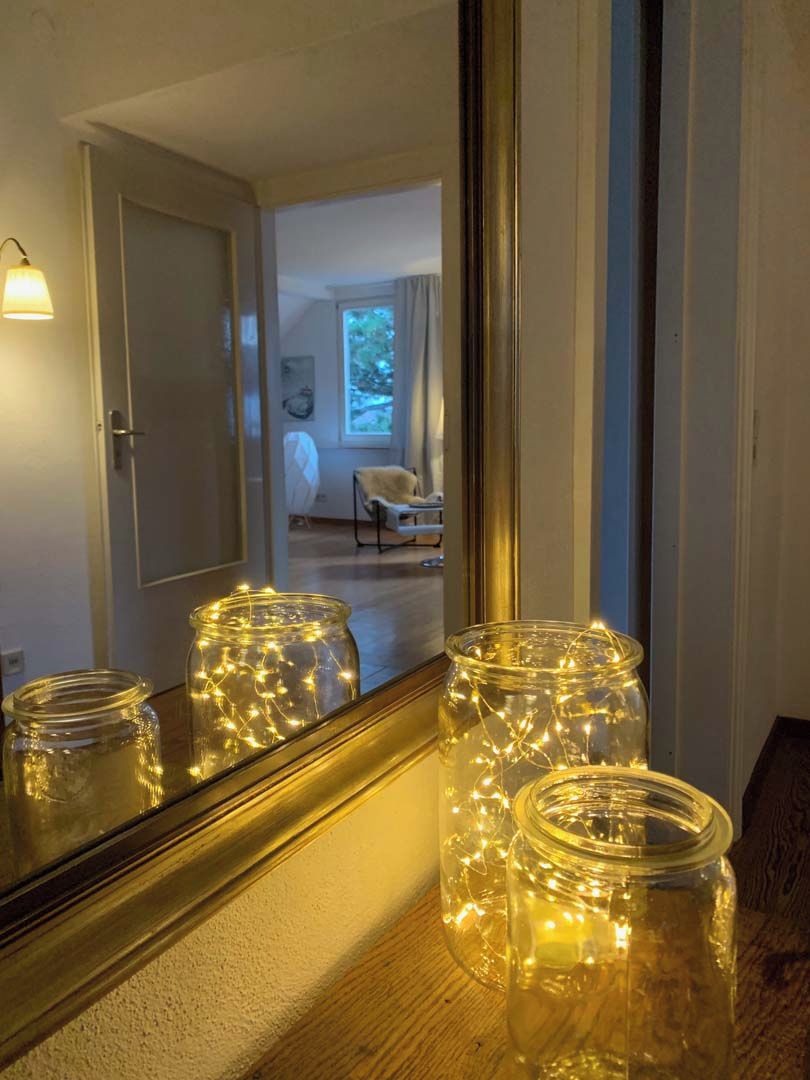 A room with a large mirror reflecting a door and a window. There are three glass jars with string lights on a wooden surface.