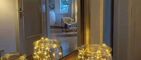 A room with a large mirror reflecting a door and a window. There are three glass jars with string lights on a wooden surface.