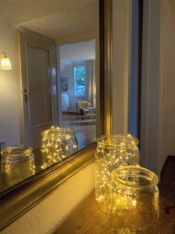 Une pièce avec un grand miroir qui reflète une porte et une fenêtre. On y voit trois bocaux en verre, décorés de guirlandes lumineuses, posés sur une surface en bois.