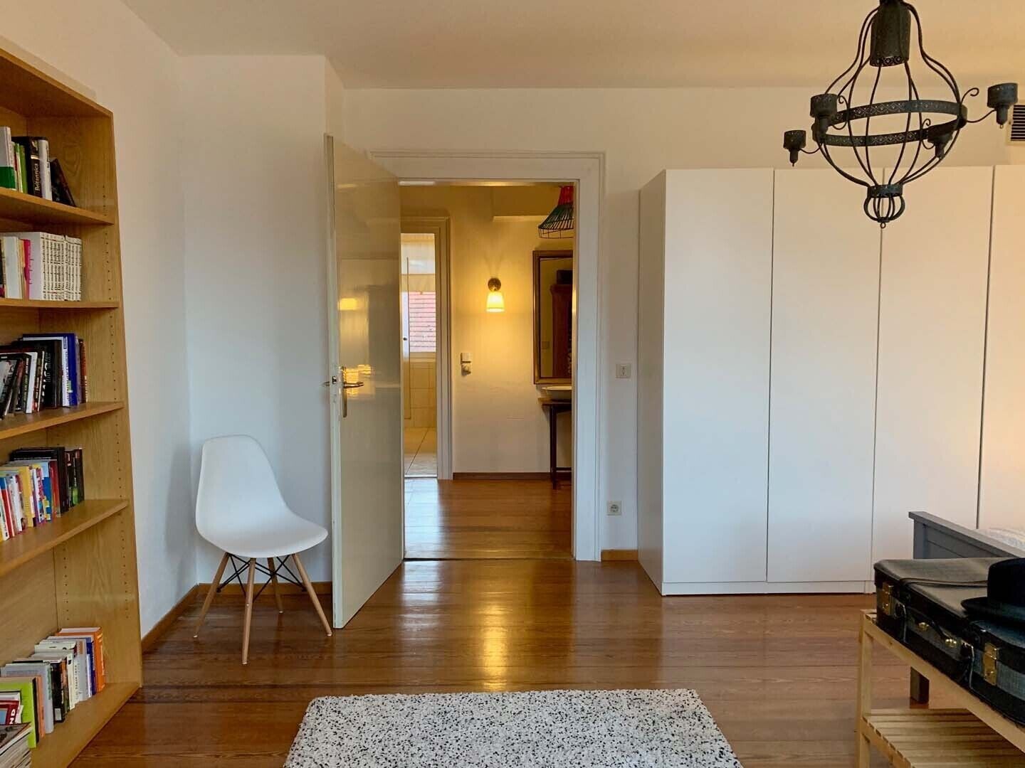 A modern living room with a wooden floor, a white chair, a bookshelf, a small rug, and a suitcase.