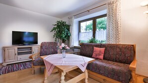 A living room with a sofa, a table, a television, and a large window with curtains. - Apartment with stunning mountain views - dream vacation in the mountains (Schönau am Königssee)