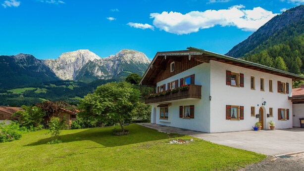 A two-story building with a balcony, surrounded by a well-maintained lawn and mountains in the background. - Apartment with stunning mountain views - dream vacation in the mountains (Schönau am Königssee)