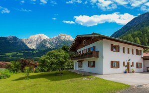 Un edificio de dos pisos con un balcón, rodeado de un césped cuidado y montañas al fondo.