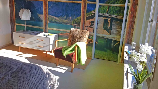 Ein Zimmer mit großem Fenster und Blick auf die Berge, einem weißen Schreibtisch mit Schubladen, einem Sessel mit grüner Decke und einer kleinen Topfpflanze.