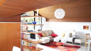 Ein modernes Wohnzimmer mit Holzbalkendecke, einem weißen Sofa, einem roten und orangefarbenen Teppich und einem Bücherregal voller Bücher.