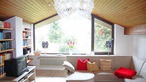 Ein modernes Wohnzimmer mit einer Couch, Bücherregalen, einem Fernseher und einem großen Fenster mit Blick auf Grünflächen.