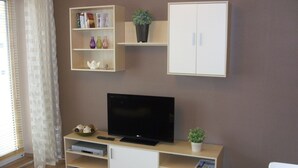 A living room with a flat-screen TV on a wooden stand, a bookshelf with books and decorative items, a cabinet, and a small potted plant. - Nice apartment near the beach and the center (Grömitz)