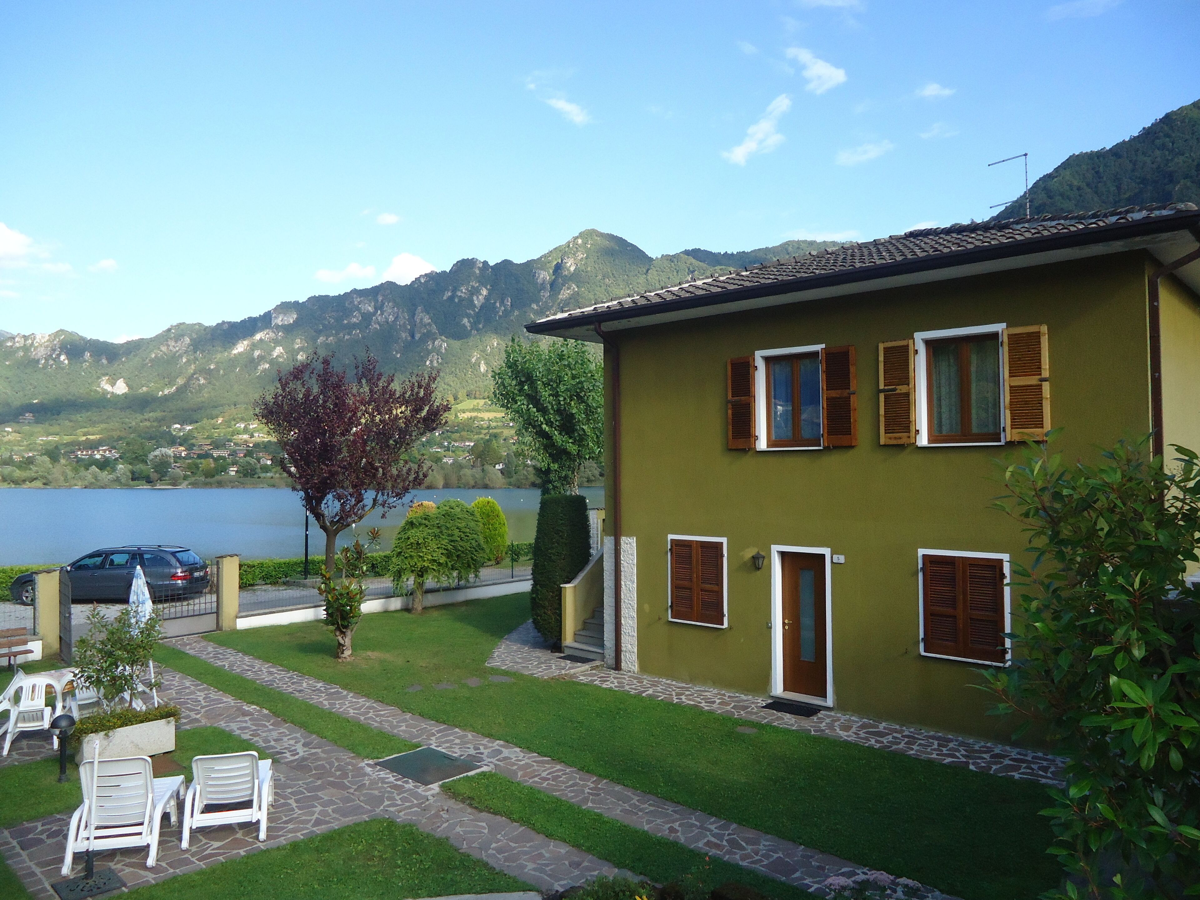 Ein Haus mit einem gepflasterten Weg, der zu einer Sitzgelegenheit mit Stühlen und Tisch führt, mit Blick auf einen See und Berge.