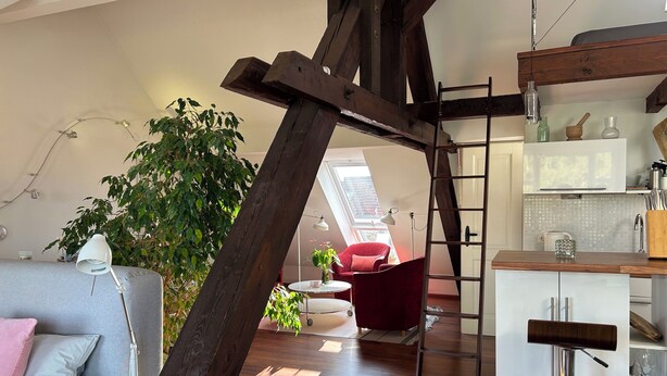A loft-style living space with a wooden beam structure, a red chair, a white sofa, and a kitchen area with a bar stool. - Attic studio in the Geteviertel - close to the city, trade fair, university, stadium and Weser cycle path (Schwachhausen)