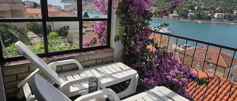 A balcony with white chairs and a table, overlooking a coastal town with boats and buildings.