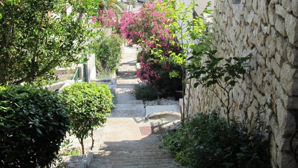 Una escala de pedra que condueix a un edifici amb teulades vermelles, envoltat de vegetació abundant i flors roses.