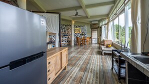 A spacious kitchen with a large refrigerator, wooden cabinets, and a stone-patterned wall. There is a dining area with chairs and a table, and a window providing a view of the outdoors. - Tirta Asri Ubud Villa (Ubud)
