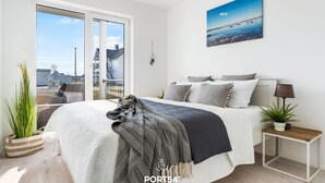 Una camera da letto con un letto, un comodino, una lampada, una pianta in vaso e vista su un edificio attraverso una porta di vetro.