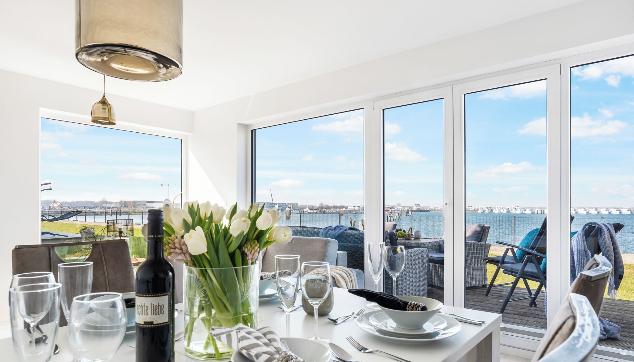 Un coin repas avec une table dressée pour deux personnes, une bouteille de vin et un vase de fleurs. De grandes fenêtres offrent une vue sur la mer et un pont.