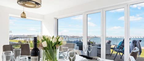 A dining area with a table set for two, a bottle of wine, and a vase of flowers. Large windows offer a view of the sea and a bridge.