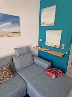 Ett modernt vardagsrum med en grå soffa, en mönstrad prydnadskudde, en röd väska och två inramade konstverk på en turkos vägg.