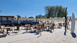 En café ved stranden med borde og stole, et surfbræt og en bygning i baggrunden.