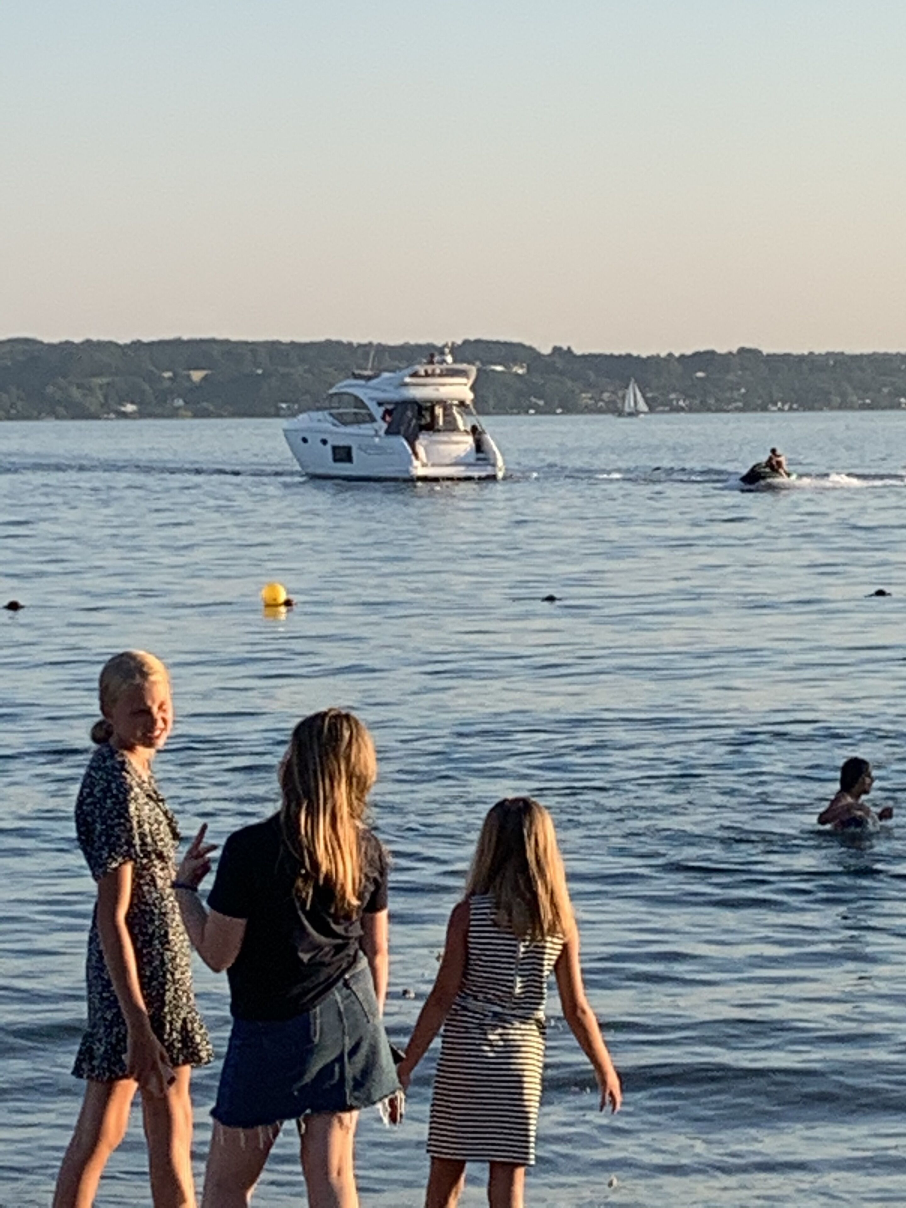 A waterfront scene with a yacht, people walking by the water, and a sailboat in the distance.