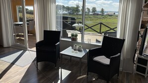 Une pièce avec deux fauteuils en osier noir, une petite table avec une lampe et une vue sur une terrasse et la verdure à l’extérieur.