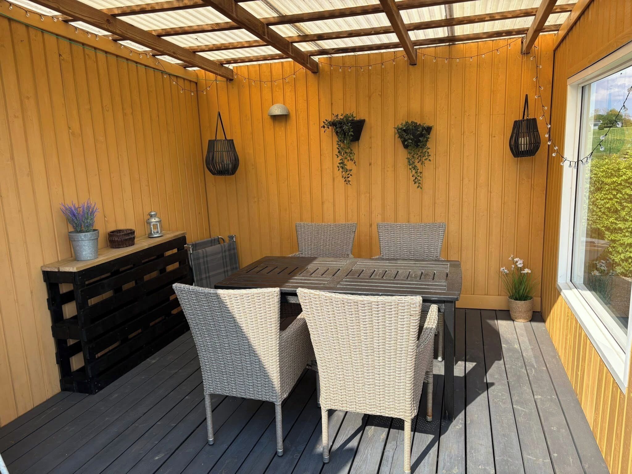 Un porche cubierto con piso de madera, una mesa con sillas, macetas colgantes y una ventana.