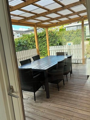 A covered patio with a wooden table and chairs, a white fence, and greenery in the background. - Holiday house with sea view (Aabenraa)
