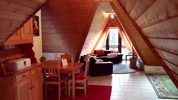 Ein Dachzimmer mit Holzbalkendecke, einem Esstisch mit Stühlen, einem Sofa und einem Fenster mit Vorhängen.