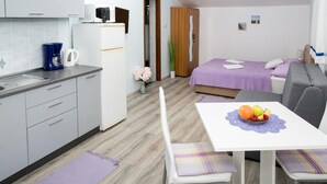 A compact hotel room with a kitchenette, a bed with a purple bedspread, a dining table with a fruit bowl, and a grey sofa.