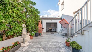 A courtyard with a stone bench, a statue, potted plants, and a view of a building with a tiled roof.