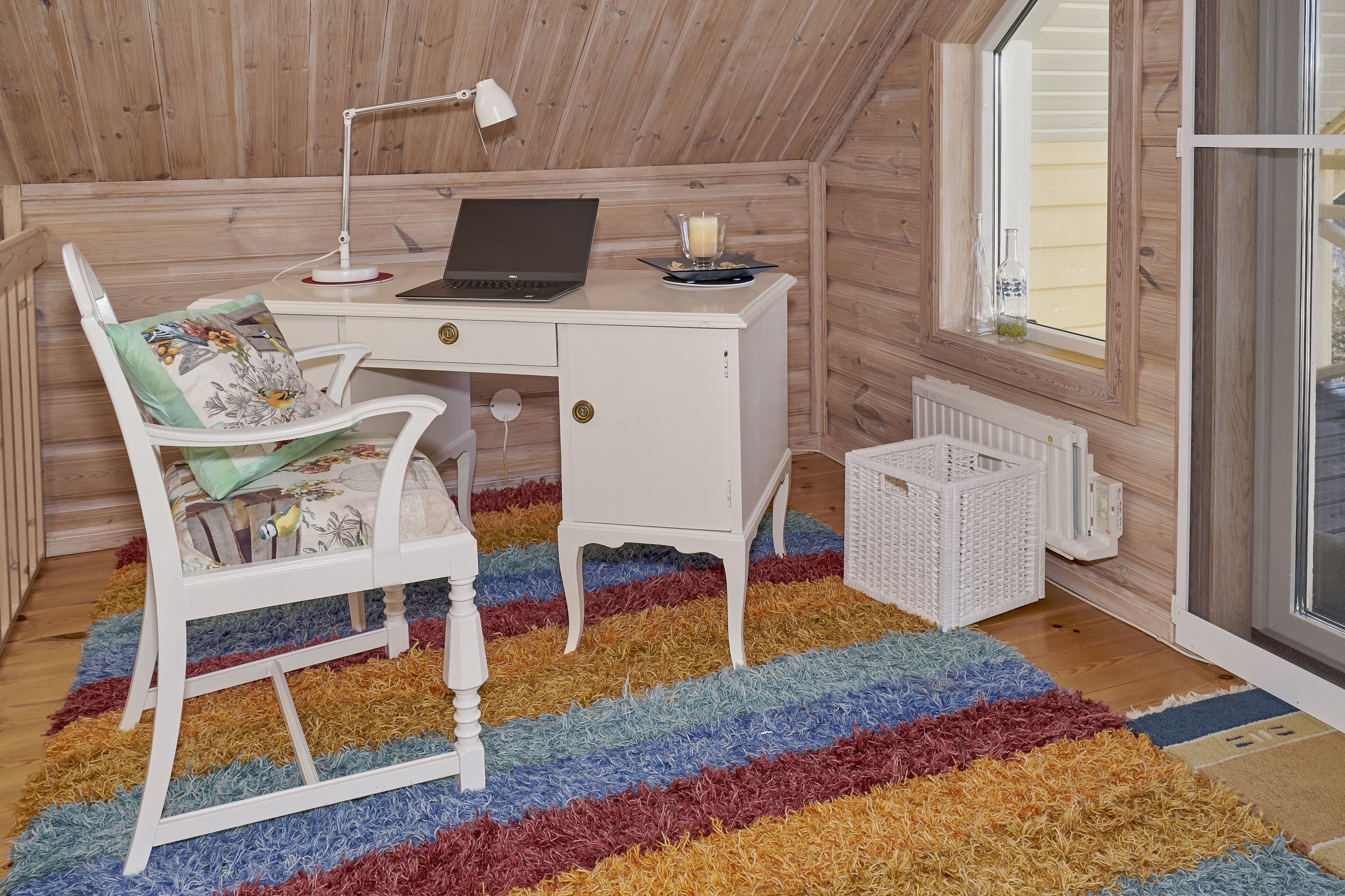 A room with a white desk, a laptop, a chair, a colorful rug, and a window with blinds.
