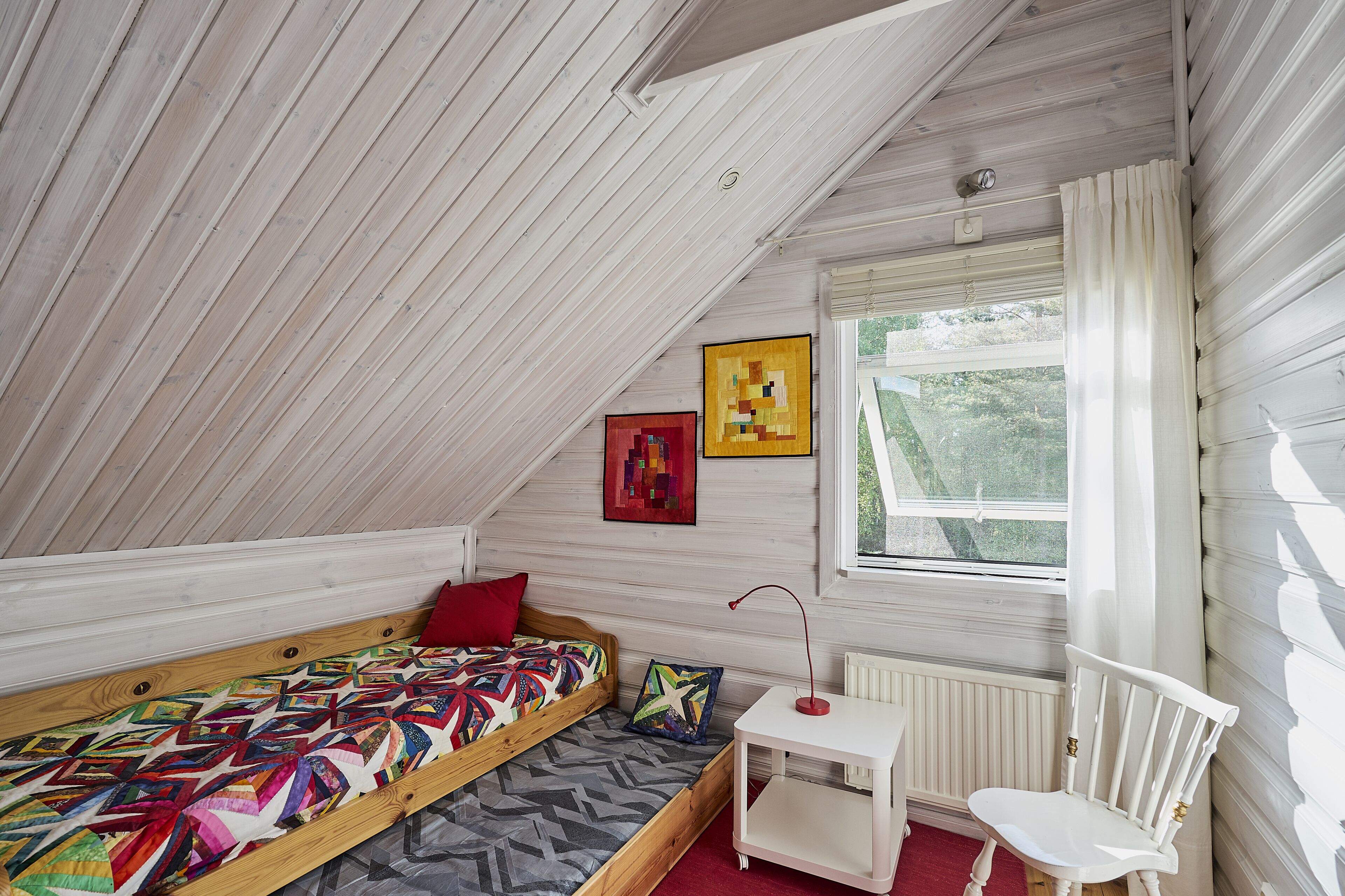 A small, cozy room with a sloped ceiling, a single bed with a colorful quilt, a small white table, a chair, and a window with curtains.