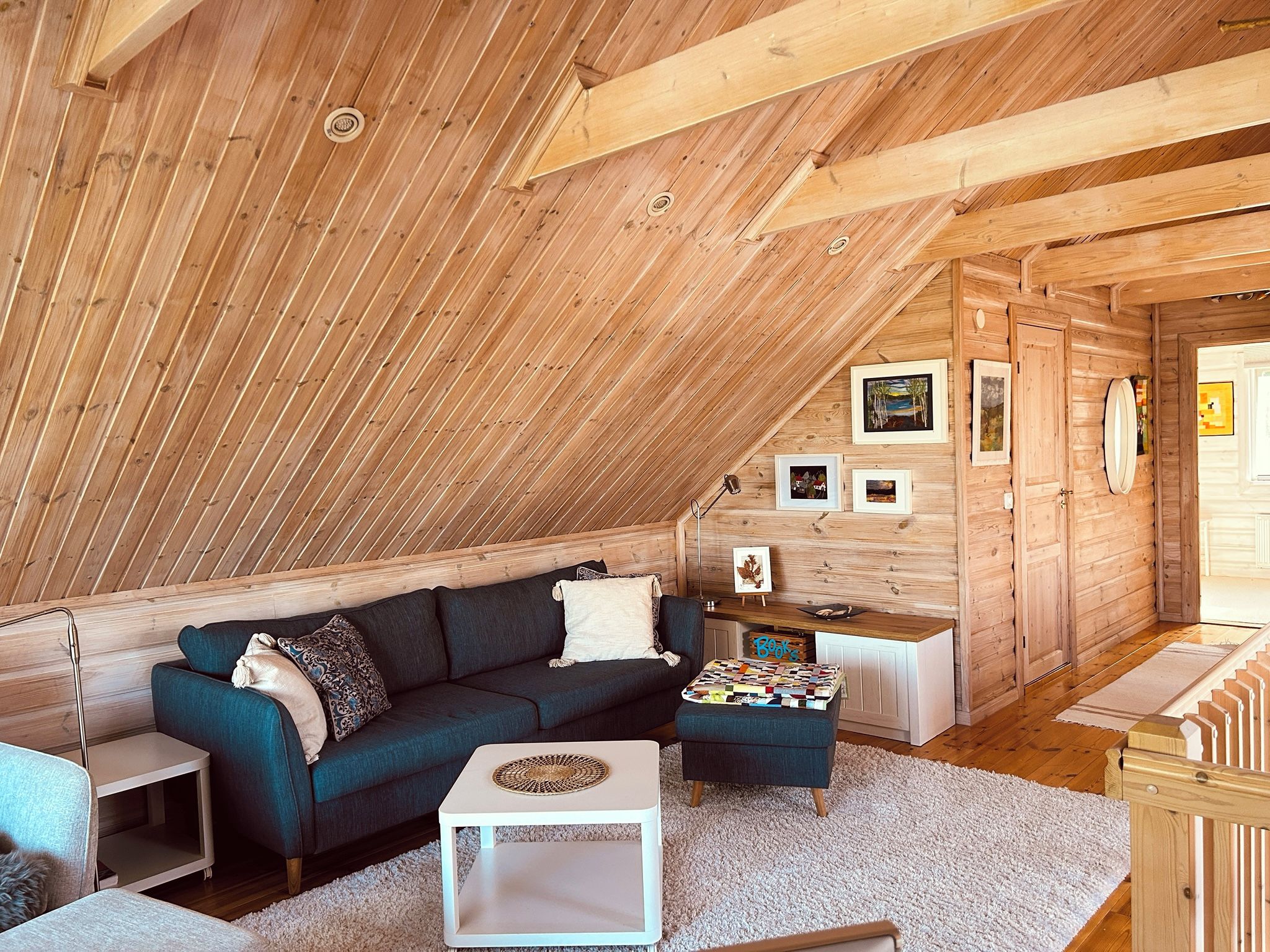 A cozy attic room with a wooden sloped ceiling, a dark blue sofa, a white side table, and a small rug.