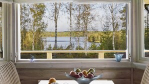A room with a large window overlooking a lake and trees, a wooden bench, and a table with a bowl of fruit. - Idyllic lakeside location, luxury furnishings - sauna, boat, canoe, WLAN, 3 bedrooms. (Nybro S)