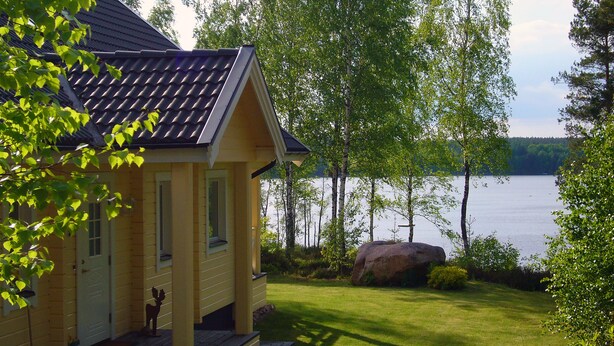A yellow house with a dark roof, a porch, and a view of a lake. - Idyllic lakeside location, luxury furnishings - sauna, boat, canoe, WLAN, 3 bedrooms. (Nybro S)