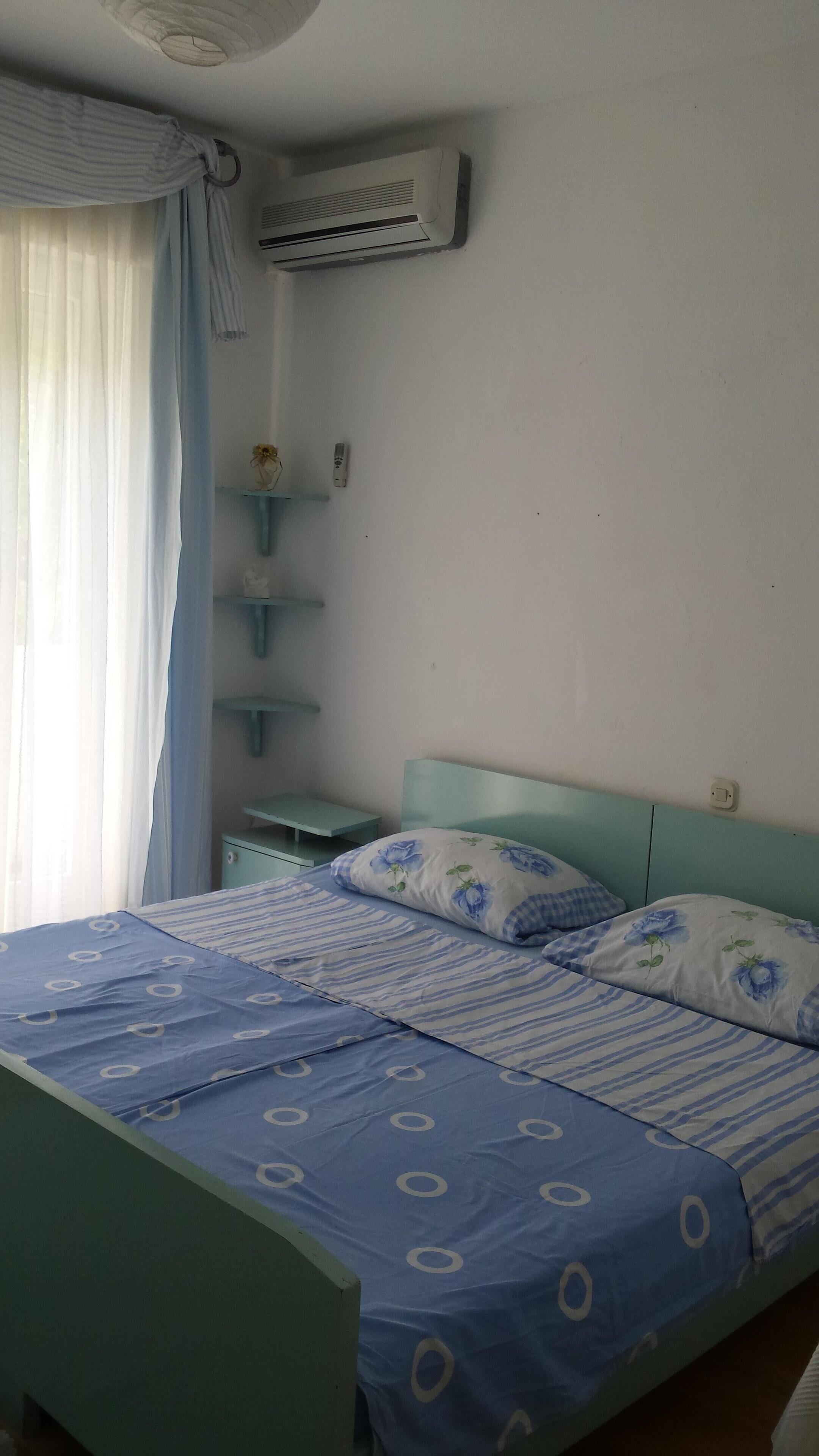 Een slaapkamer met een bed, nachtkastje, een luchtverkoeler aan de muur en een raam met gordijnen.