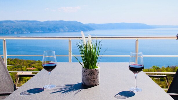 A table with two wine glasses and a potted plant, overlooking a scenic view of a body of water and distant hills. - Holiday house with fantastic sea view and pool - spring offer from 27. 4.-19. 5th (Ripenda Kosi)