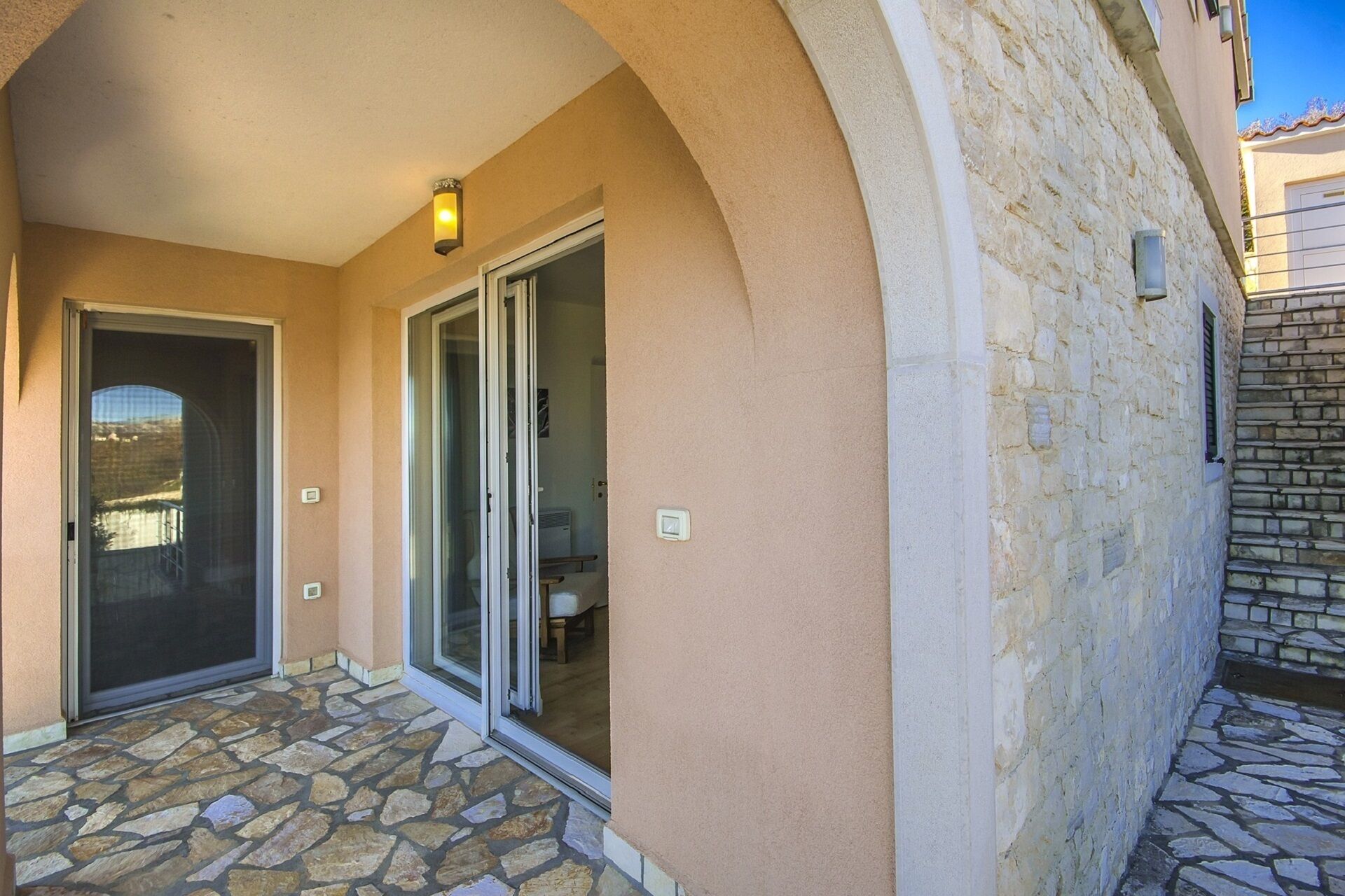 A building entrance with a stone-tiled porch, a sliding glass door, and a small light fixture above the door.