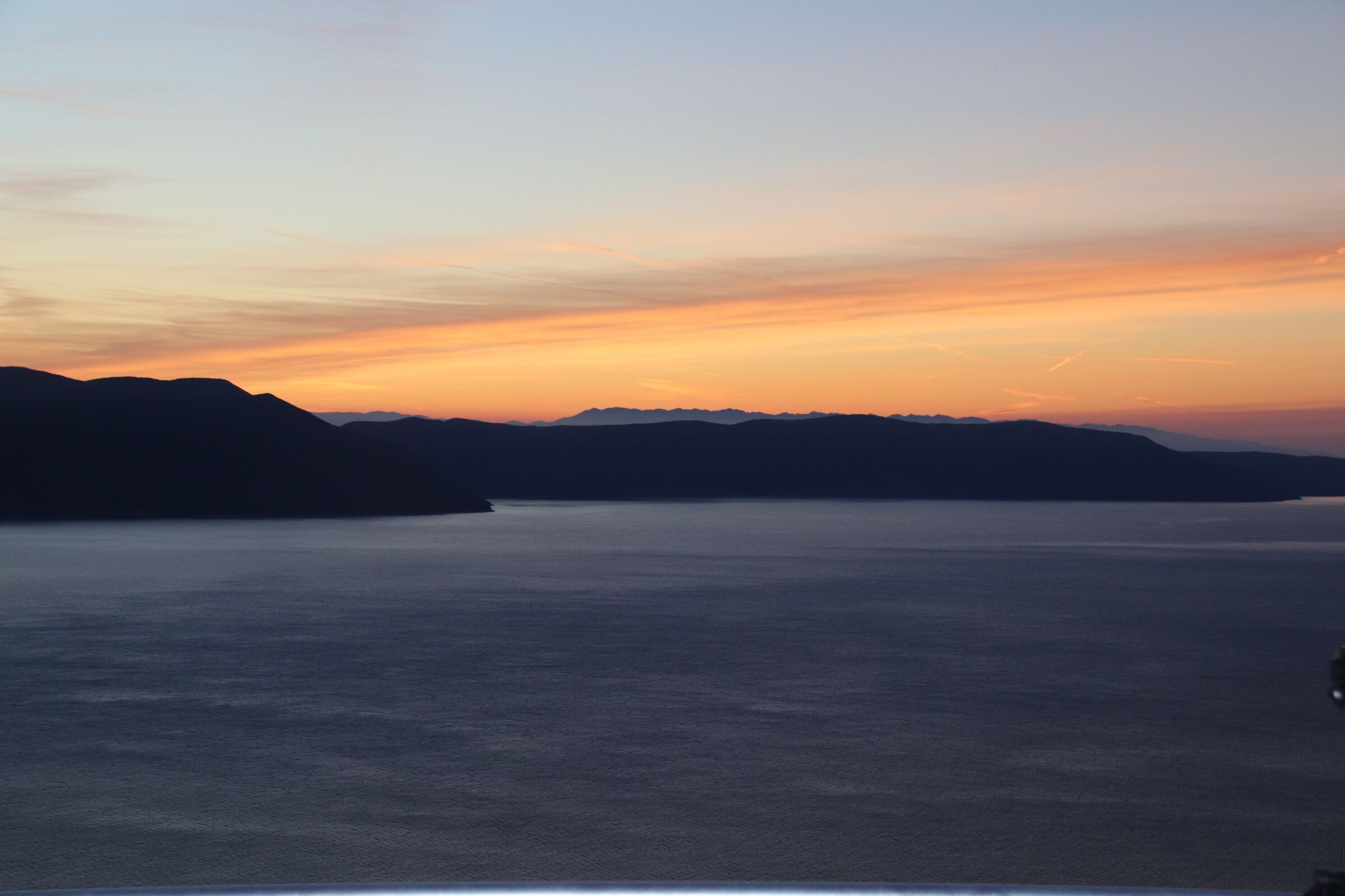 A coastal landscape with a calm sea and distant mountains during sunset.