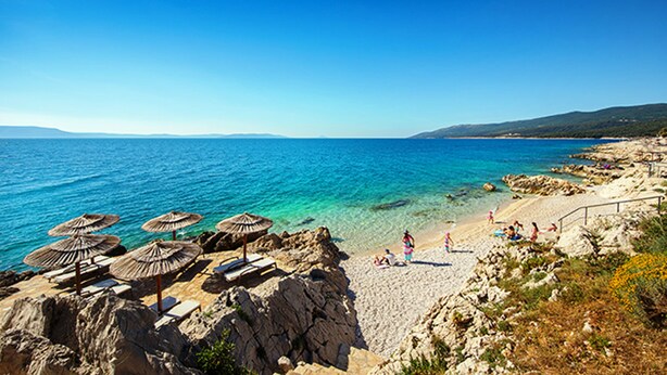 A beach with clear blue water, rocky shoreline, and umbrellas.