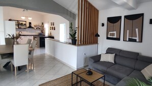 A modern living room with a grey sofa, a coffee table, and a dining area with a white table and chairs. There is a kitchen in the background with white cabinets and a black countertop.