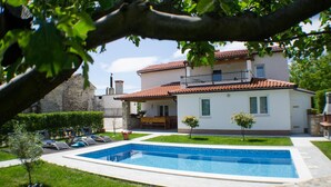 A two-story house with a swimming pool, a balcony, and a stone wall in the background.