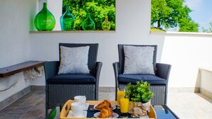 Two wicker chairs with cushions, a tray with croissants, a glass of orange juice, and a potted plant on a balcony with a view of trees.