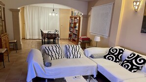 A living room with two sofas, a coffee table with remotes, a dining area with a table and chairs, and a bookshelf.