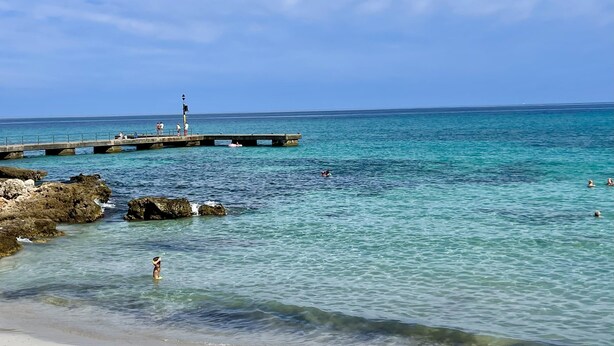 A beach with clear turquoise water, a pier extending into the sea, and rocky outcrops near the shore.