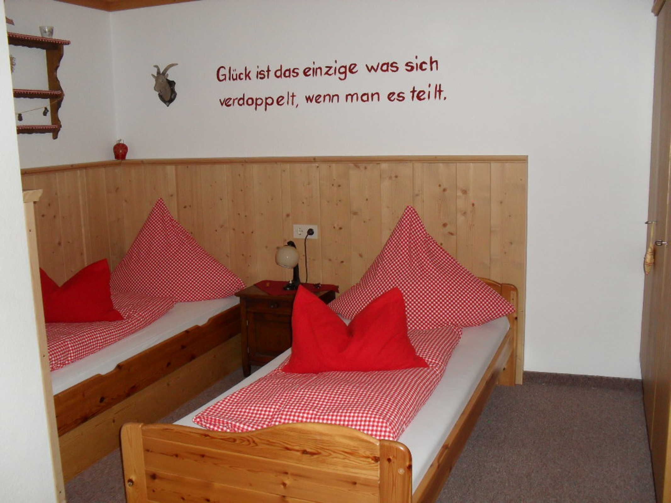 A room with two wooden bunk beds, each with a red and white checkered bedspread and red pillows. There is a wall-mounted shelf with decorative items and a wall with a German quote.