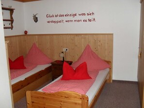 A room with two wooden bunk beds, each with a red and white checkered bedspread and red pillows. There is a wall-mounted shelf with decorative items and a wall with a German quote.