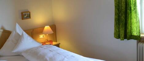 A neatly made bed with white linens, a bedside lamp, a framed picture on the wall, and a window with green curtains.
