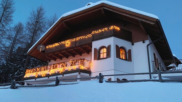 Ein schneegebdecktes Chalet mit einem Balkon, der mit Lichterketten, Holzbalken und einer weißen Fassade verziert ist.