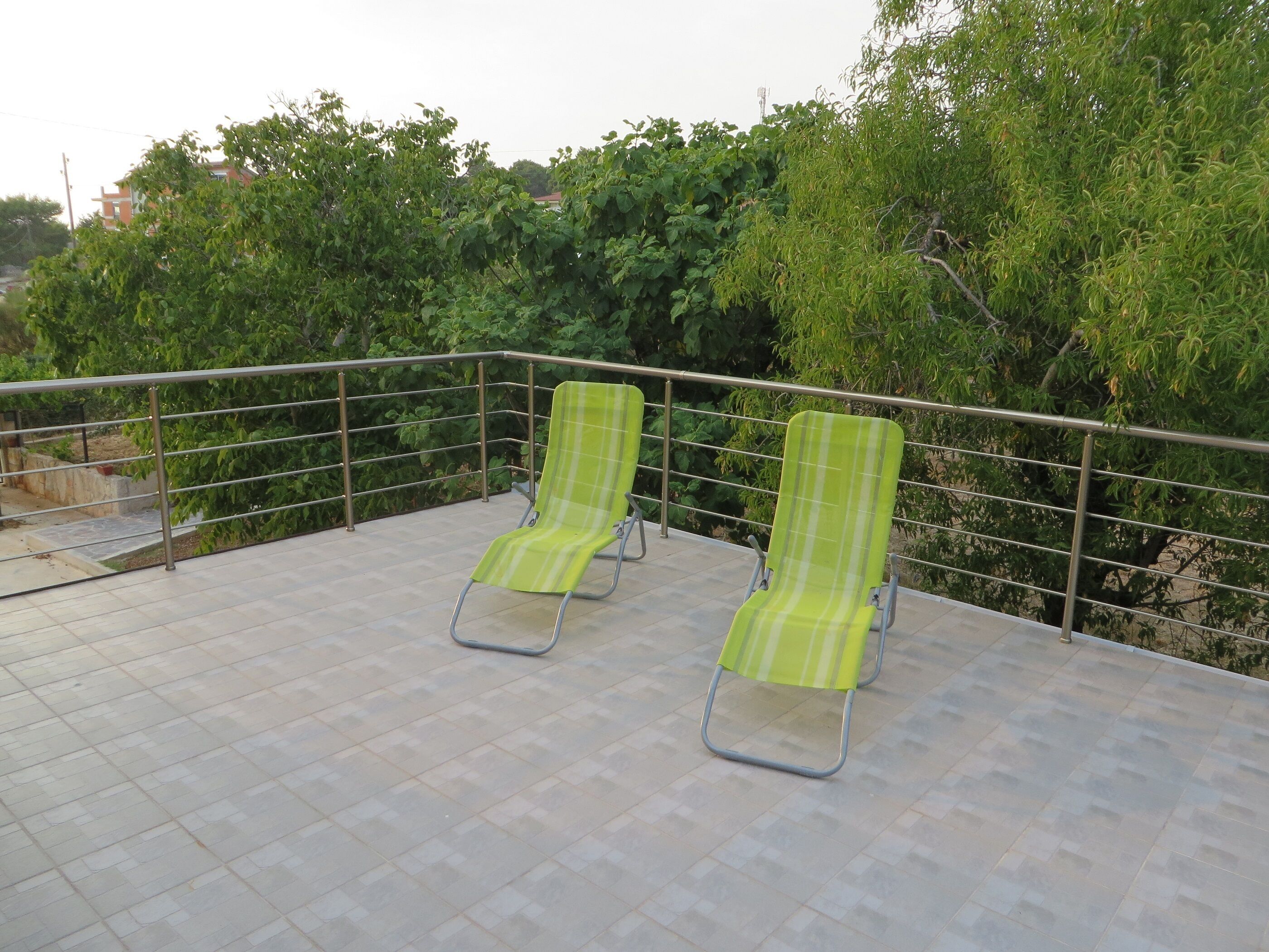 Deux fauteuils de jardin à rayures vertes, placés sur une terrasse carrelée et bordée d’une rambarde en métal, le tout entouré d’une végétation luxuriante.