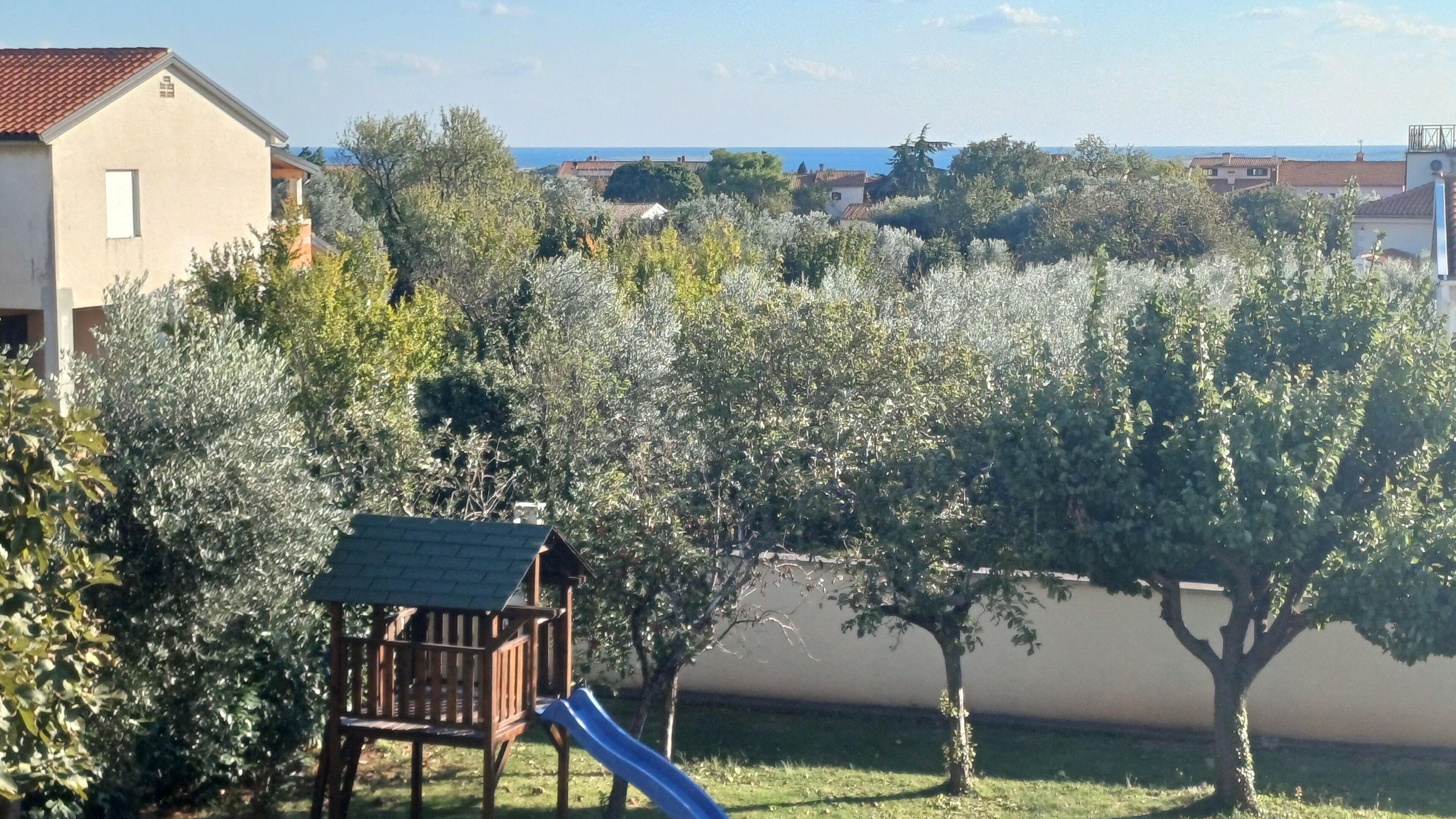 Ein Spielplatz mit Rutsche und einem hölzernen Spielhäuschen, umgeben von Olivenbäumen und Blick aufs Meer.
